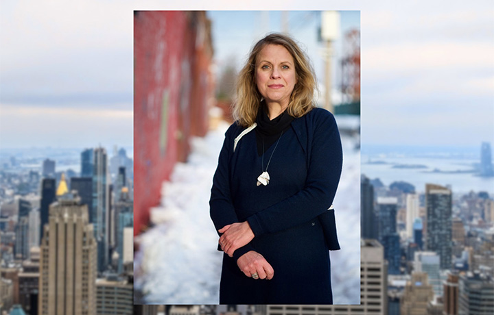 Color photo of a person in a black coat, with a blurred photo of the New York City skyline in the background
                                           