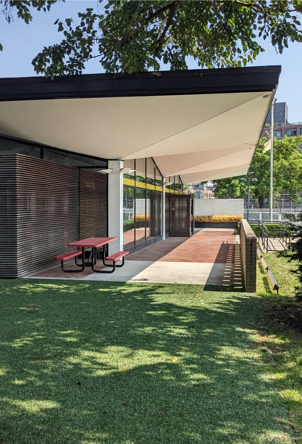 A modern single-story building with umbrella-like roof extending beyond exterior walls, surrounded by a green lawn. Picnic table visible on building’s patio, mid-rise residential building visible in the background.