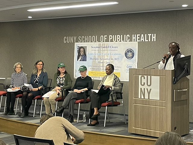 New York State Senator Cordell Cleare speaking at a podium, with other panelists seated to her right. Signage above the seated panelists reads “CUNY School of Public Health” and a poster contains information about the forum, including the date and time.