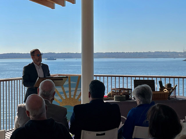 Man at podium speaking to people seated in front of him, standing in front of artwork and marine artifacts, with body of water and ship in the background 