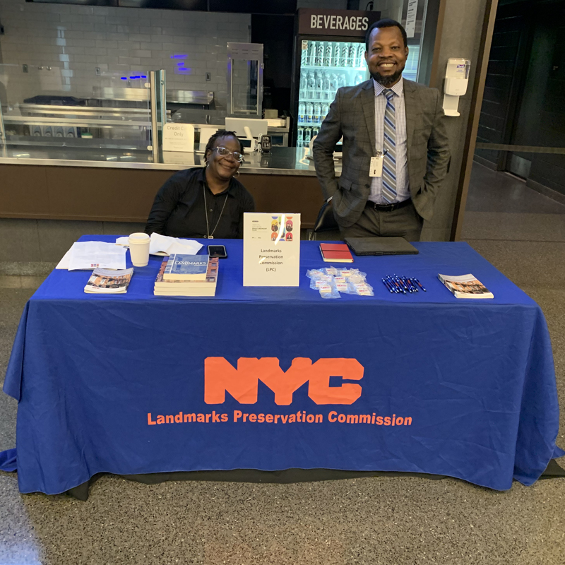 Two smiling people behind a table with covering reading NYC Landmarks Preservation Commission