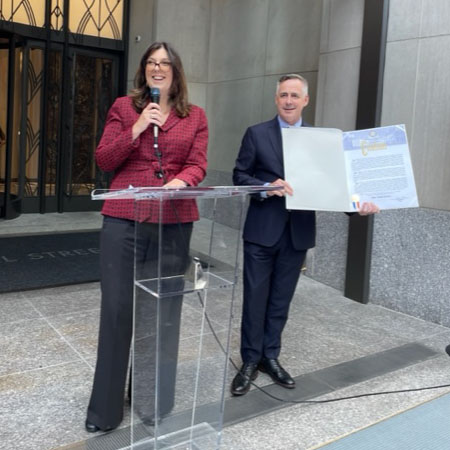 LPC Chair Sarah Carroll stands with a microphone at a podium outside a building entrance, and NYLPF Chair Tom Krizmanic stands next to her holding the Mayoral Citation