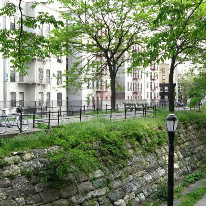 A public walkway with benches and a parked bicycle on top of a stone wall, with a streetlamp in the foreground and buildings lining the far side of the walkway