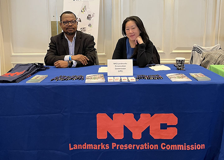 Two people sitting behind a table covered with pamphlets and a blue tablecloth with the logo reading NYC Landmarks Preservation Commission