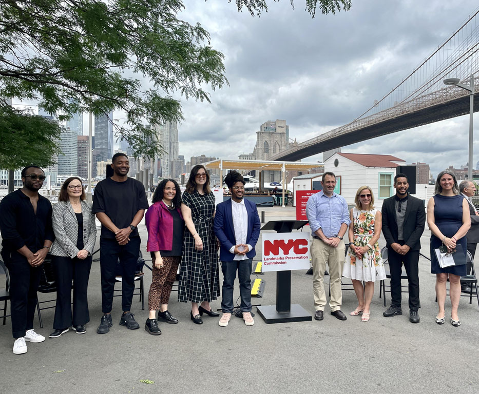 Group of smiling people standing around podium with Brooklyn Bridge and NYC skyline in background 