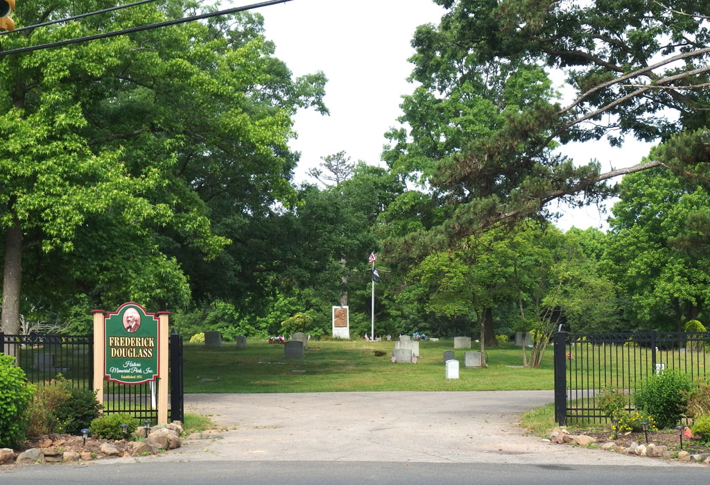 Entrance to fenced cemetery with gravestones, memorial, and flagpole, sign reads 'Frederick Douglass Historic Memorial Park