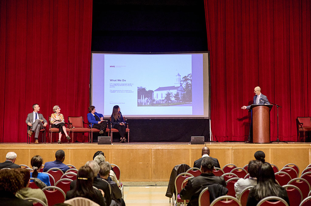 Four people seated on a stage in front of a screen, one person standing at podium to speak