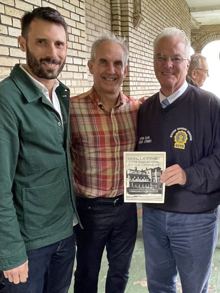 Three smiling people standing in front of a tan brick wall and holding a piece of paper that reads Living Landmarks, A Tour of Historic Homes in St. George, Staten Island