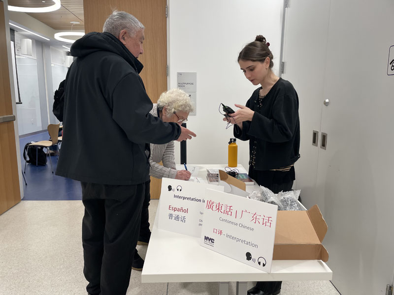 Three people standing by a table with signs reading Interpretation – Español and Interpretation – Cantonese Chinese in their respective languages. One person is programming a headset for listening.