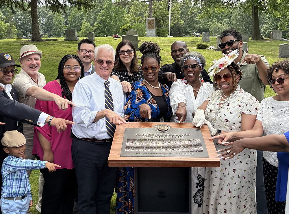 Photo of smiling people pointing at a historic plaque; cemetery and headstones in the background