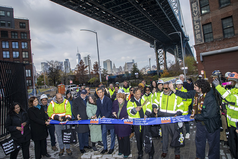 Color image of group of smiling people cutting a ribbon with Manhattan bridge in the background, interior of historic building with tall windows and statue in the righthand corner
