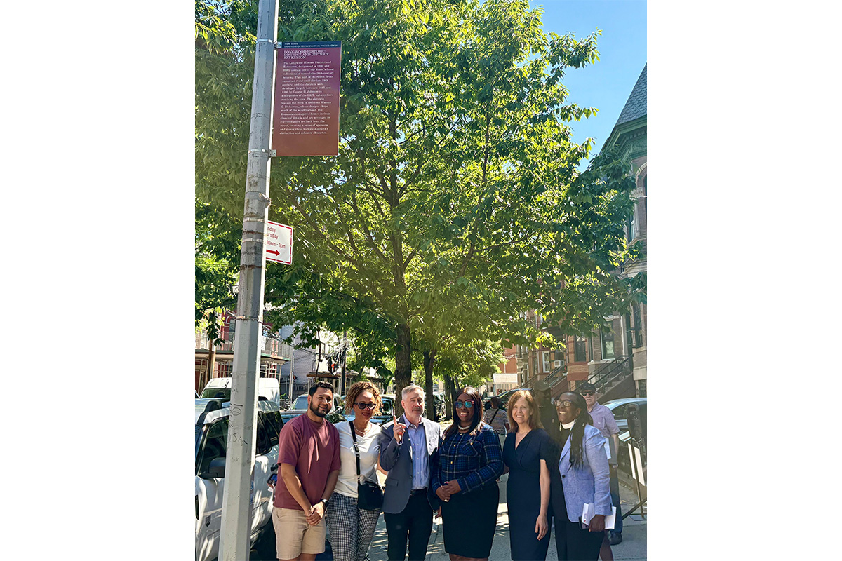 Color photo of smiling people standing beneath a historic marker mounted on a street pole, with historic homes in the background