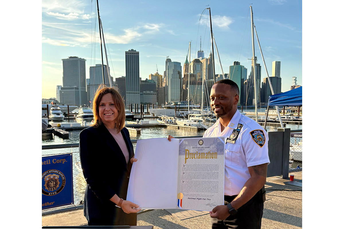 Color photo of two smiling people holding a large mayoral plaque with boats, the river, and Manhattan's skyline in the background