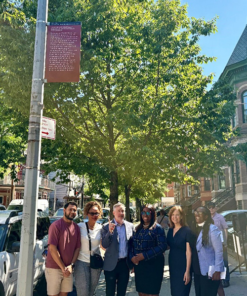 Smiling people standing next to a pole with red sign attached at the top and historic row homes in the background