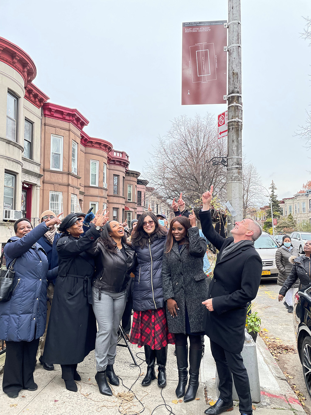 New York City Landmarks Preservation Commission (LPC) Chair Sarah Carroll, New York Landmarks Preservation Foundation (NYLPF) Chair Tom Krizmanic, Council Member Farah Louis and residents of the East 25th Street in East Flatbush are looking up and point at the historic district marker attatched to a lamp post