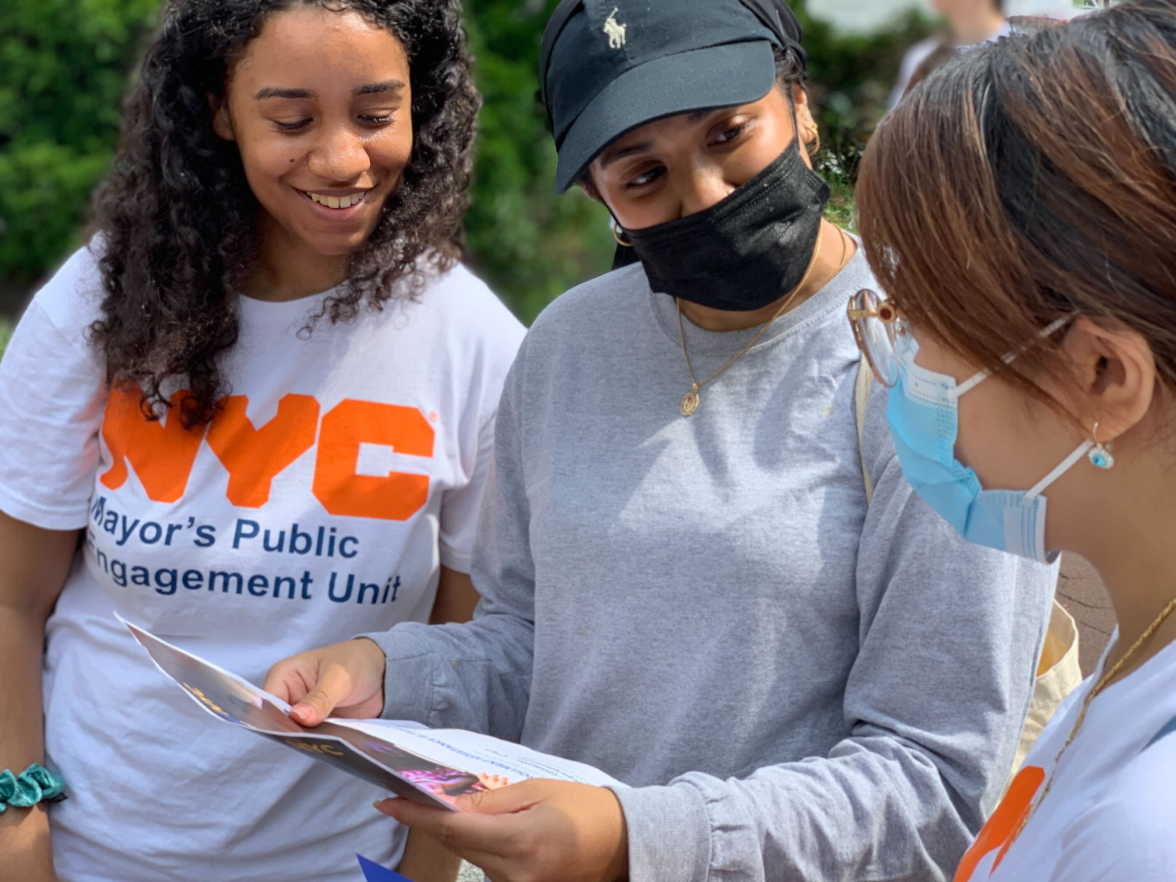 Two CUNY interns wearing PEU T-shirts talk to a woman wearing a hat about a flyer she was given. 