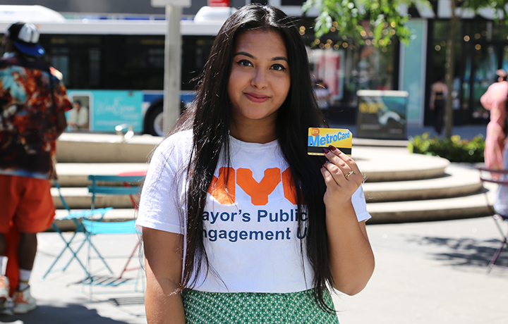A person wearing a PEU t-shirt smiles at the camera and holds up a MetroCard in her left hand.