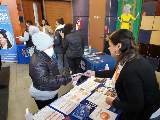 PEU staff stands behind a table filled with flyers as she hands a flyer to another person who wears a winter hat and grabs the flyer. Other people can be seen in the background near other tables.