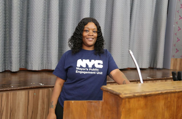 A woman stands, smiling, at aa podium, wearing a blue NYC Mayor's Public Engagement Unit tee-shirt.