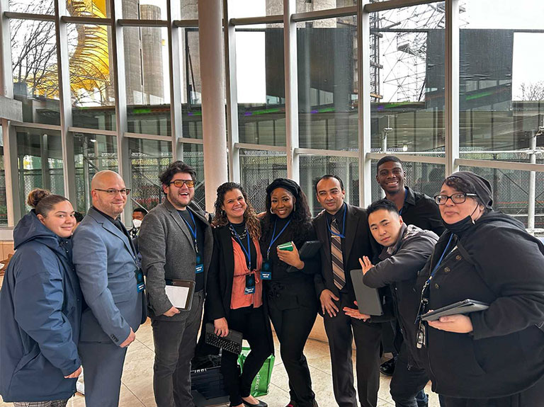 A Group of PEU staffers in business casual dress stand in a group and pose together inside in font of big windows, all smiling.