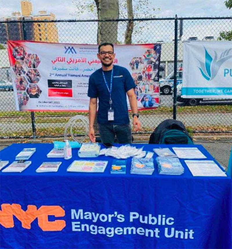 A man stands behind a table, smiling, wearing aa GetCoveredNYC tee shirt. The table reads N Y C Mayor's Public Engagement Unit and holds a lot of flyers