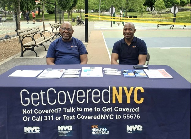 Two GetCoveredNYC Specialists sit at a Get Covered NYC table, behind them is a park. A banner on the table reads 'Get Covered NYC: Not covered? Talk to me to Get Covered or call 311 or text CoveredNYC to 55676'