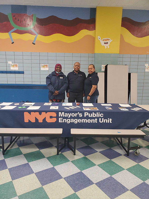 Three PEU Specialists stand behind a table smiling at the camera. Table cloth reads NYC Mayor's Public Engagement Unit and holds lots of flyers