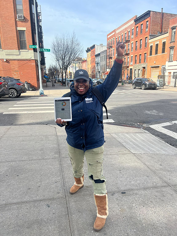 A TSU Specialist stands on the sidewalk with her fist raised, smiling in celebration, and holding up a tablet