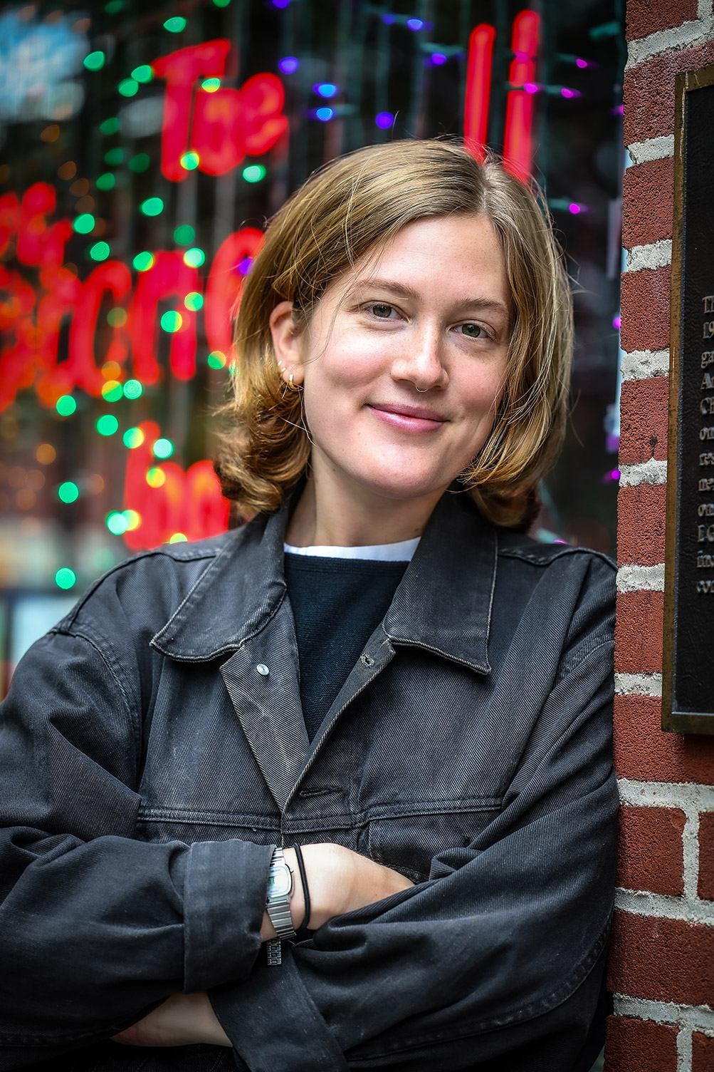 A photo of Elise smiling with her arms crossed, wearing a black denim jacket in front of the Stonewall Inn.