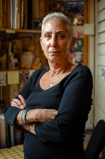 Photo of a woman in her 60s named Jill Wynn standing with her arms crossed in her home, looking into the camera. She is wearing a black long-sleeved shirt with jewelry and has short white hair.
