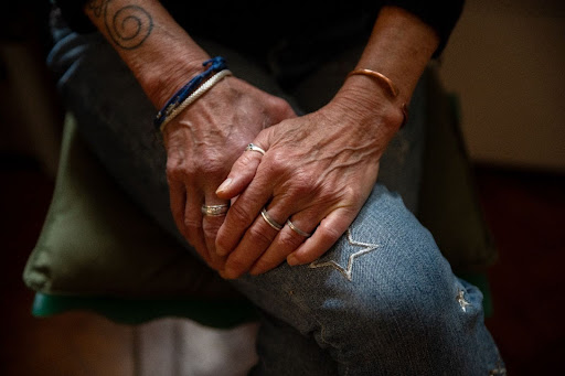 Close up photo of Jills hands resting on top of her knees. She is wearing rings and bracelets and jeans with star designs on them. A tattoo is showing on her arm.