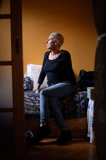 Photo of Jill sitting on her bed with her legs crossed and looking to the side. Her profile is lit by natural light from a window she is looking out of outside the frame.