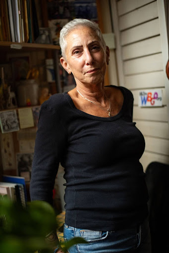 Photo of Jill looking straight into the camera with a soft smile. Her arms are resting by her side. She is in her home with a bookshelf in the background and a plant in the foreground.