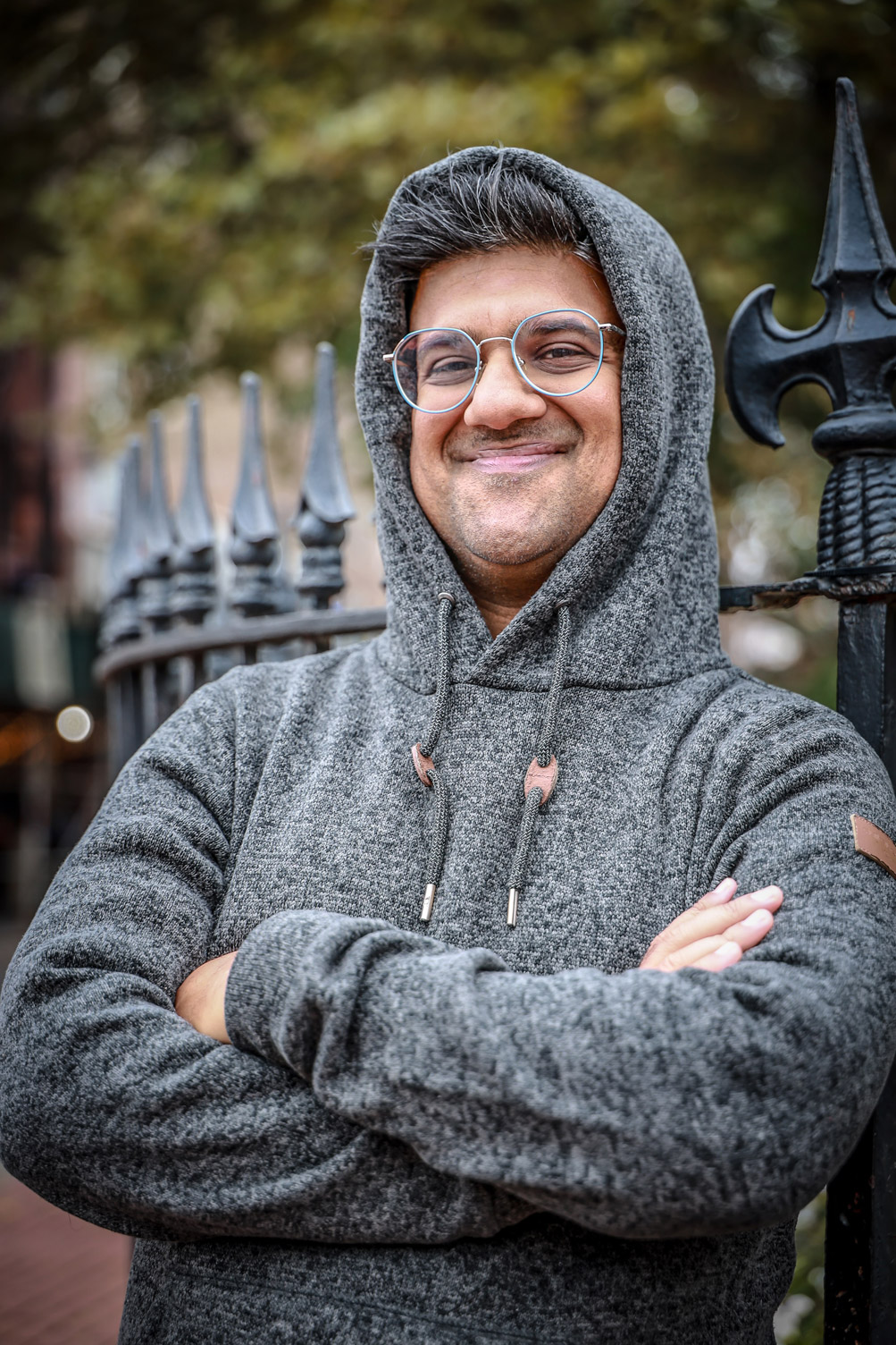 A photo of Nishant smiling with his arms crossed outside in front of a gate. He has his gray hoodie on over his head.
