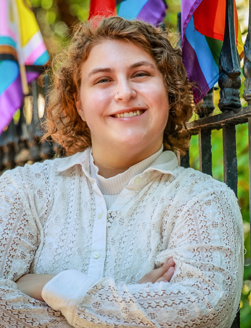 Woman with folded arms standing next to wrought iron fence displaying Pride Flags