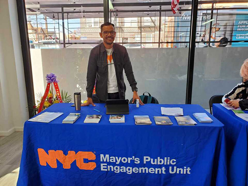 GetCoveredNYC Specialist stands at a Public Engagement Unit  table with flyers piled on top of it