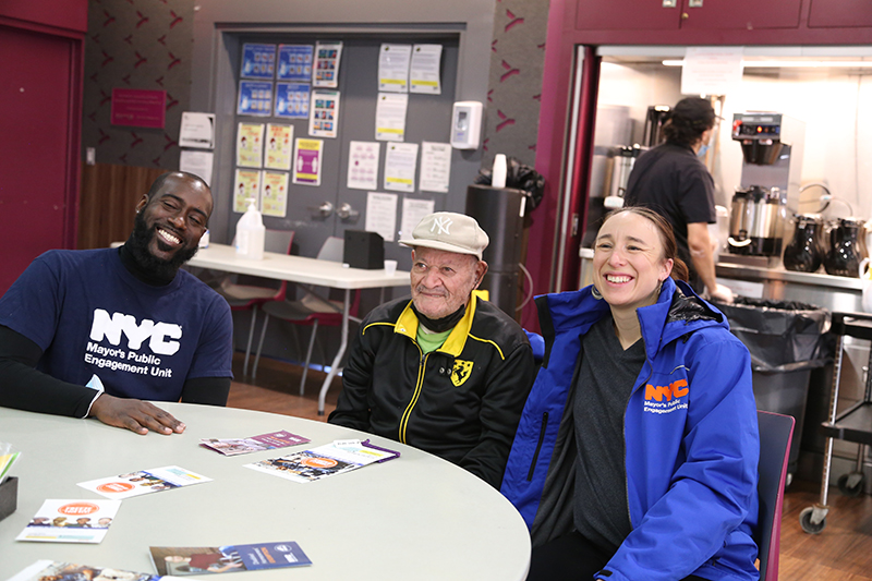 An older man sits with two  P E U staffers, with rent freeze flyers in front of them on the table