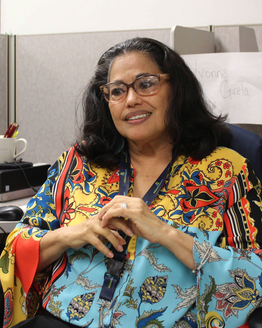 Photo of H S U Specialist, Yvonne, smiling while sitting by her desk. She has dark hair and is wearing glasses, a bright patterned blouse, and a Home Support Unit lanyard. 