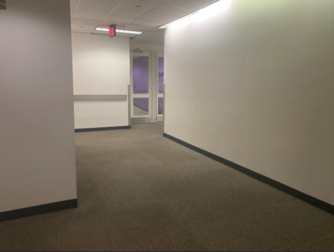 This is a hallway with white walls that leads from the elevator to the MOCS' main entrance that has two doors with glass windows.