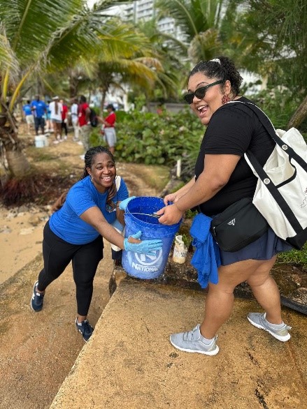 A Black woman hands a bucket of dirt to a Hispanic woman at a beach in San Juan, Puerto Rico. The women are smiling and having a great time.