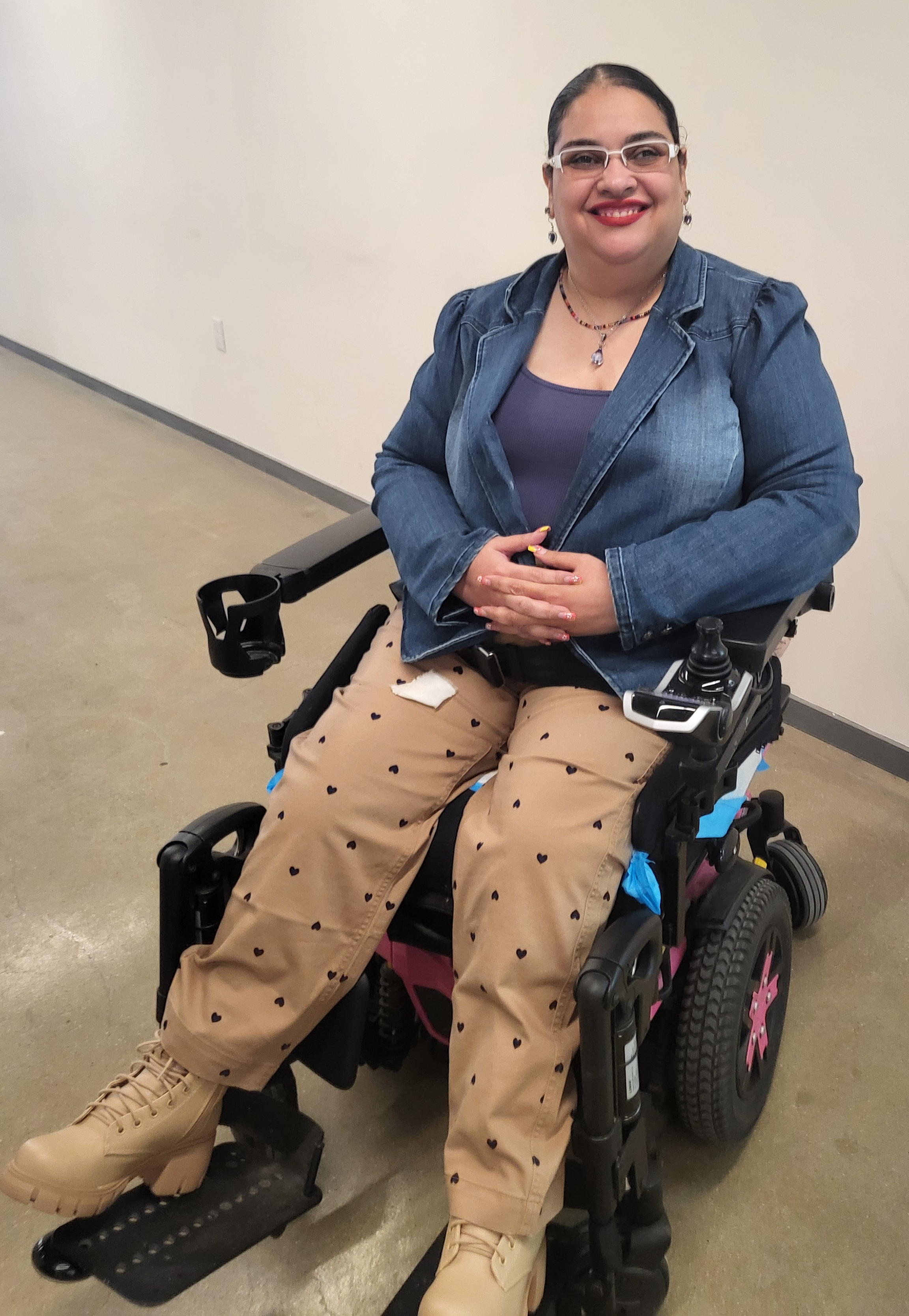 Crystal wearing glasses, a denim blazer, and tan heart-patterned pants is sitting in a motorized wheelchair with her hands folded.