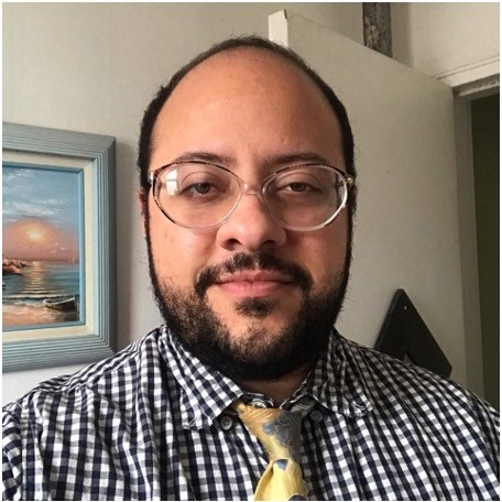 A light skinned, bald and bearded Latinx person wearing light purple glasses and a black and white checkered shirt with a yellow and blue tie. Behind them is a blue framed painting of a boat on a beach.