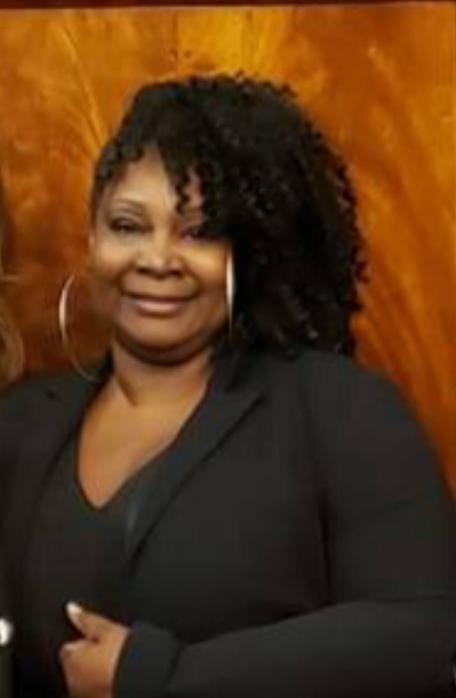 A woman of mid brown caramel skin tone wearing a black blazer and top and large hoop earrings with a gentle smile. Her curly shoulder length hair is worn to one side and is shaved low on the other. She stands in front of a warm brown paneled wood wall with one hand resting near her waist.