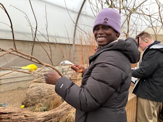 Rain wearing a black jacket and purple hat, smiling at the camera while holding two budgie birds.