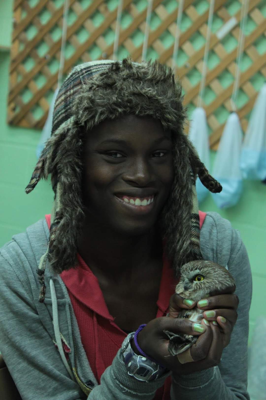 Rain wearing a hat, smiling at the camera and holding a saw-whet owl.