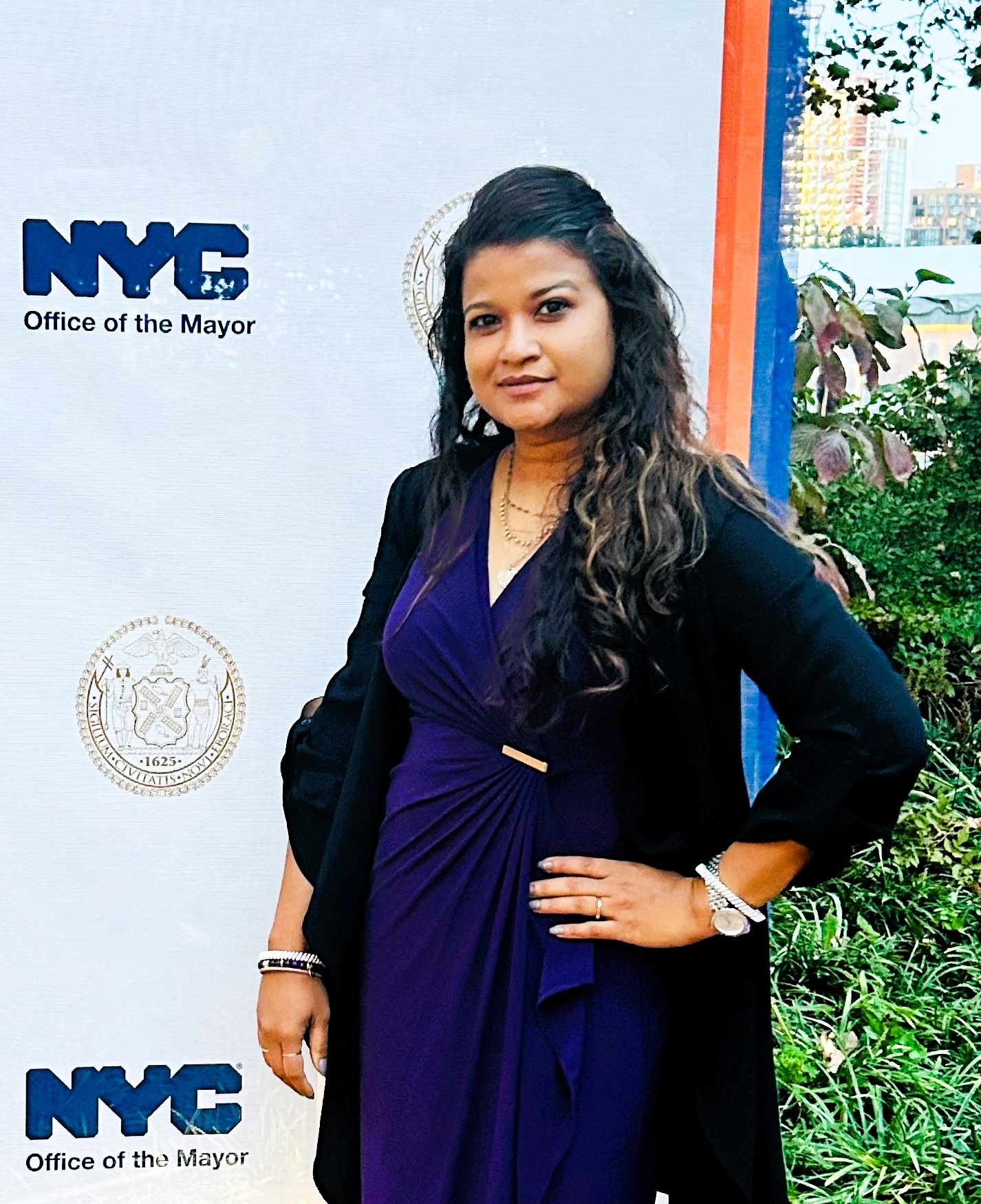 Woman with long dark hair, wearing a blue dress, standing with her hand on her hip in front of a step and repeat from a Mayor’s Office event.