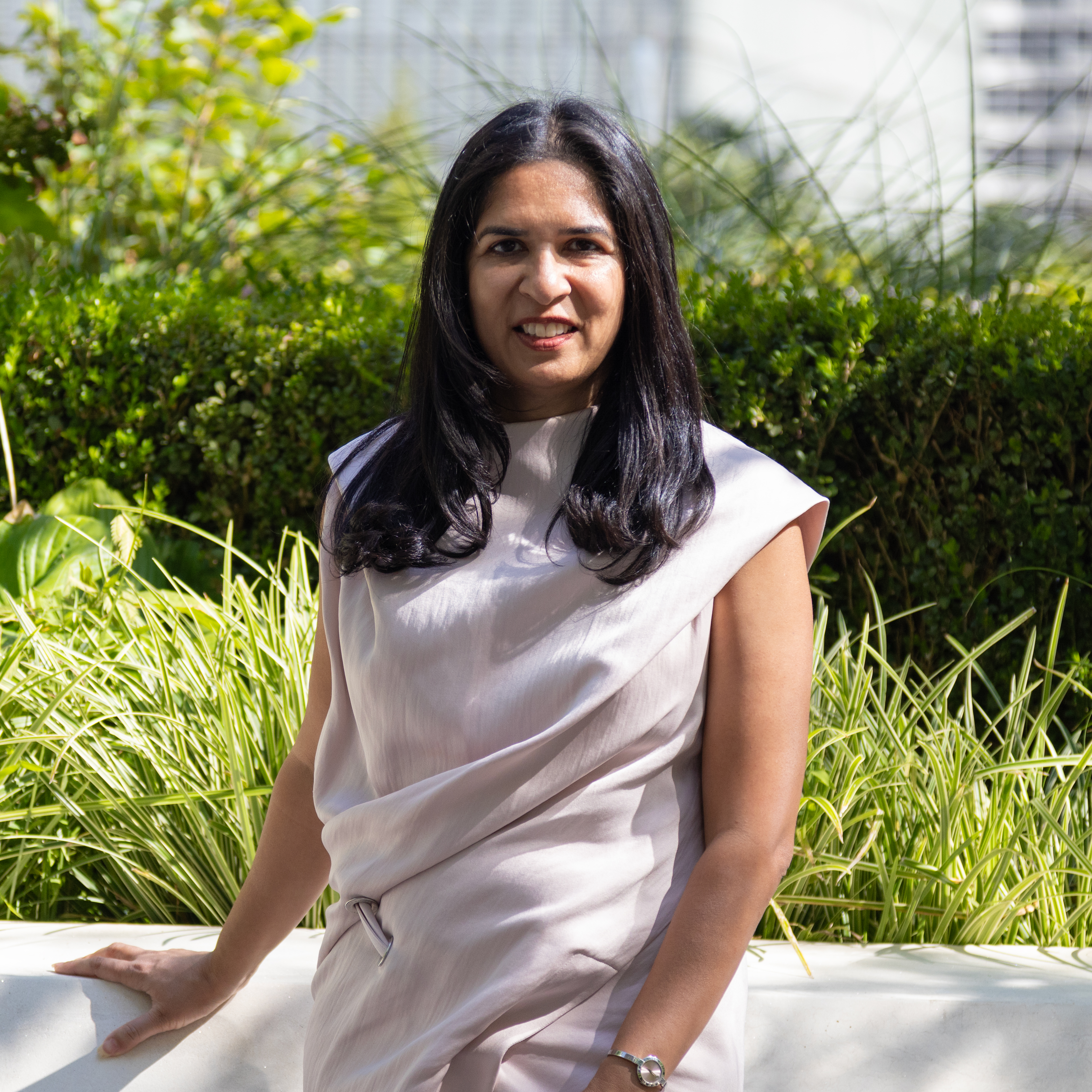 Seema has long black hair and is smiling while wearing a light-colored dress standing outdoors with lush greenery and a softly blurred landscape. 