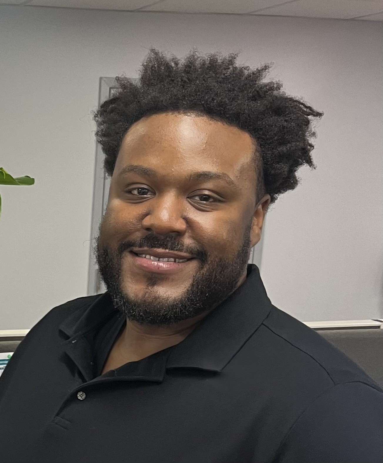 Headshot of Zachary Taylor, a dark, brown-skinned man with curly black hair, smiling in a black long-sleeve pique polo shirt.