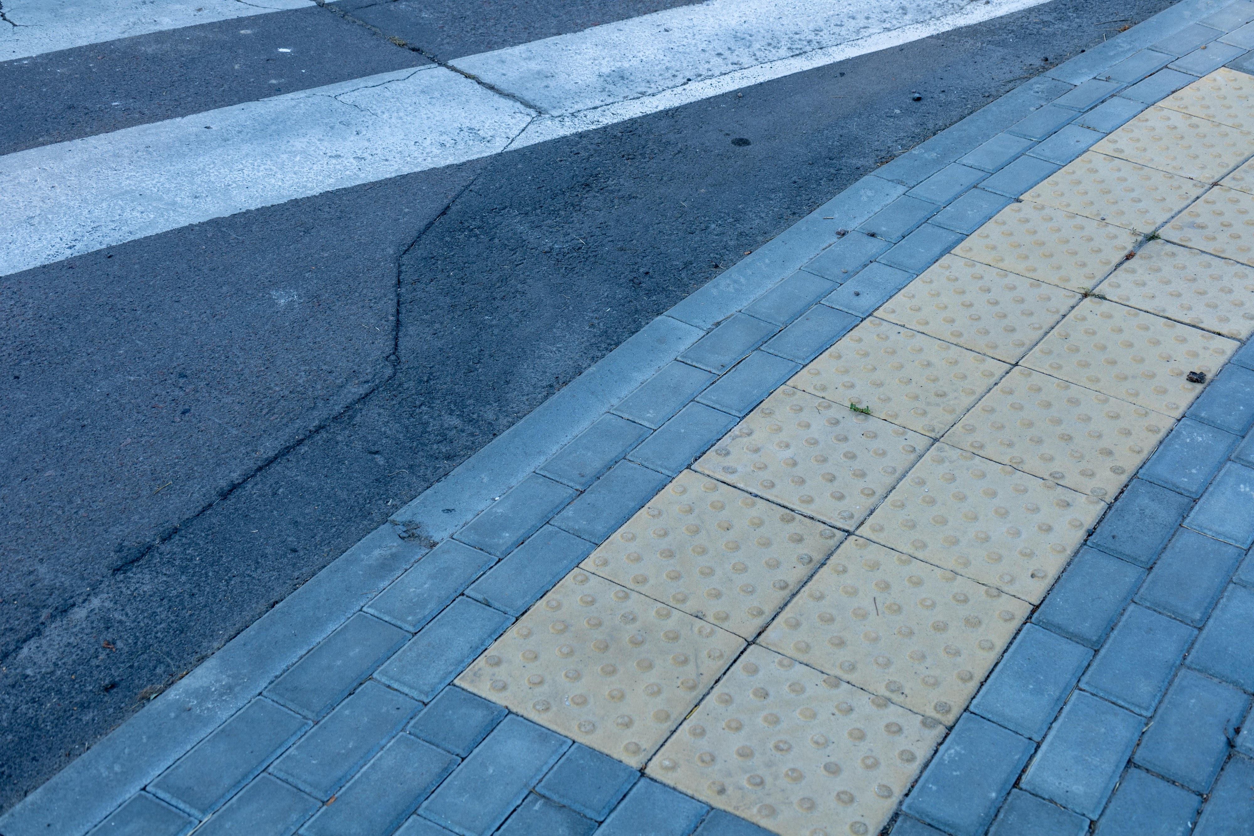 A close-up, ground-level view of yellow tactile paving tiles at a crosswalk. The tiles feature raised, flat-topped domes, also known as truncated domes, designed to be detectable underfoot.