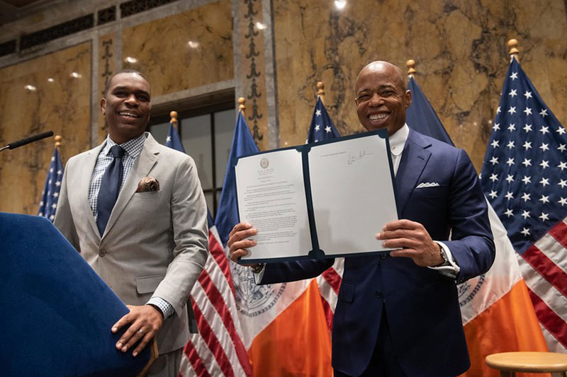 Mayor Adams stands at the podium with the Office of Faith-Based and Community Partnerships Director, Gilford Monrose. Mayor Adams is holding an Executive Order.